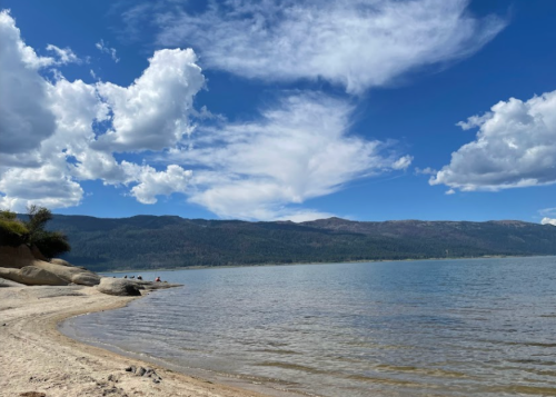 A serene lakeside view with sandy shore, calm water, and fluffy clouds against a blue sky and distant mountains.